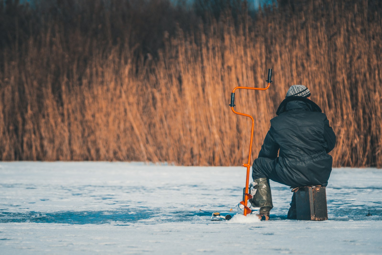 Pilkkijä istuu jäällä jääkaira vierellään, taustalla ruovikkoa